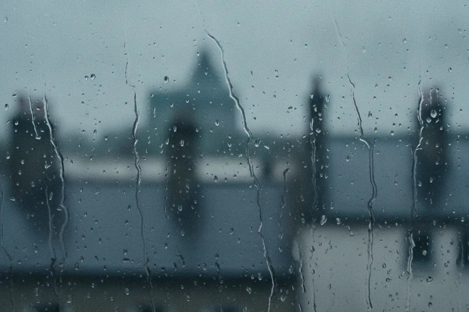 Regentropfen auf einer Fensterscheibe, dahinter unscharf ein grauer Himmel über Hausdächern