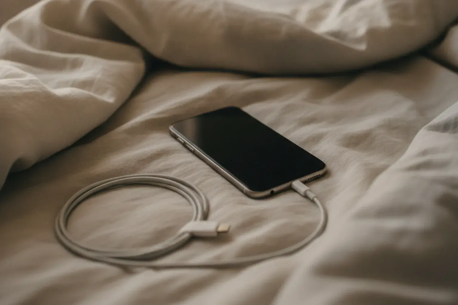 Ein Smartphone mit dunklem Bildschirm liegt auf einer Bettdecke neben einem Ladekabel