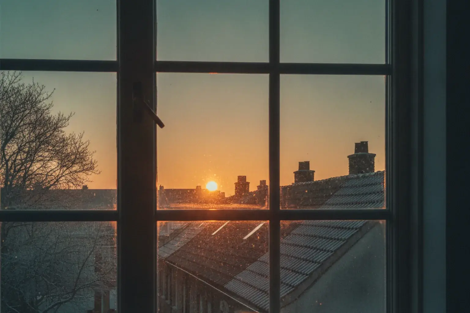 Blick aus einem Fenster auf die ersten Sonnenstrahlen über Dächern, im Vordergrund eine Tasse auf der Fensterbank
