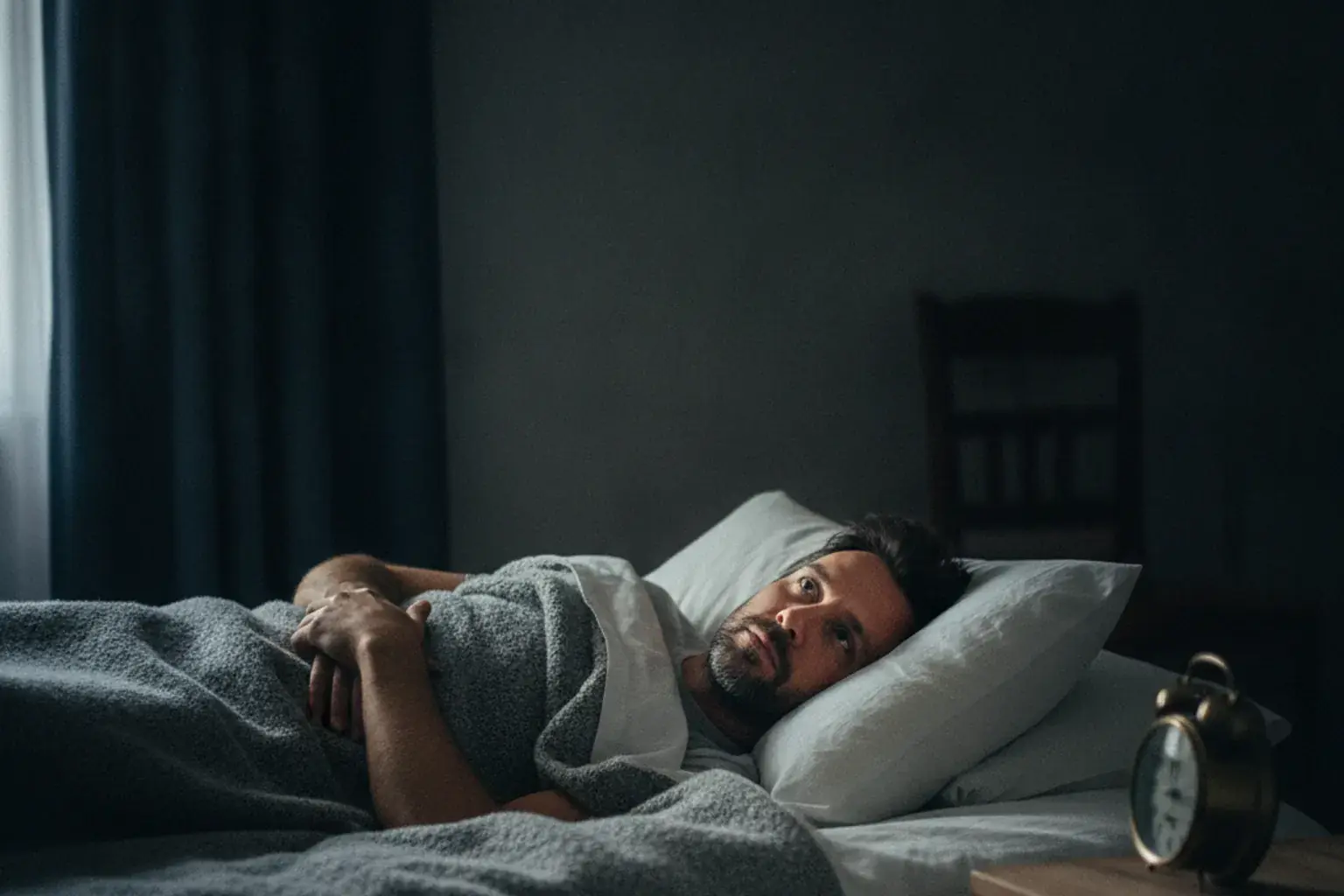 Mann liegt nachts wach im Bett und blickt ruhig zur Decke, der Wecker auf dem Nachttisch ist von ihm weggedreht